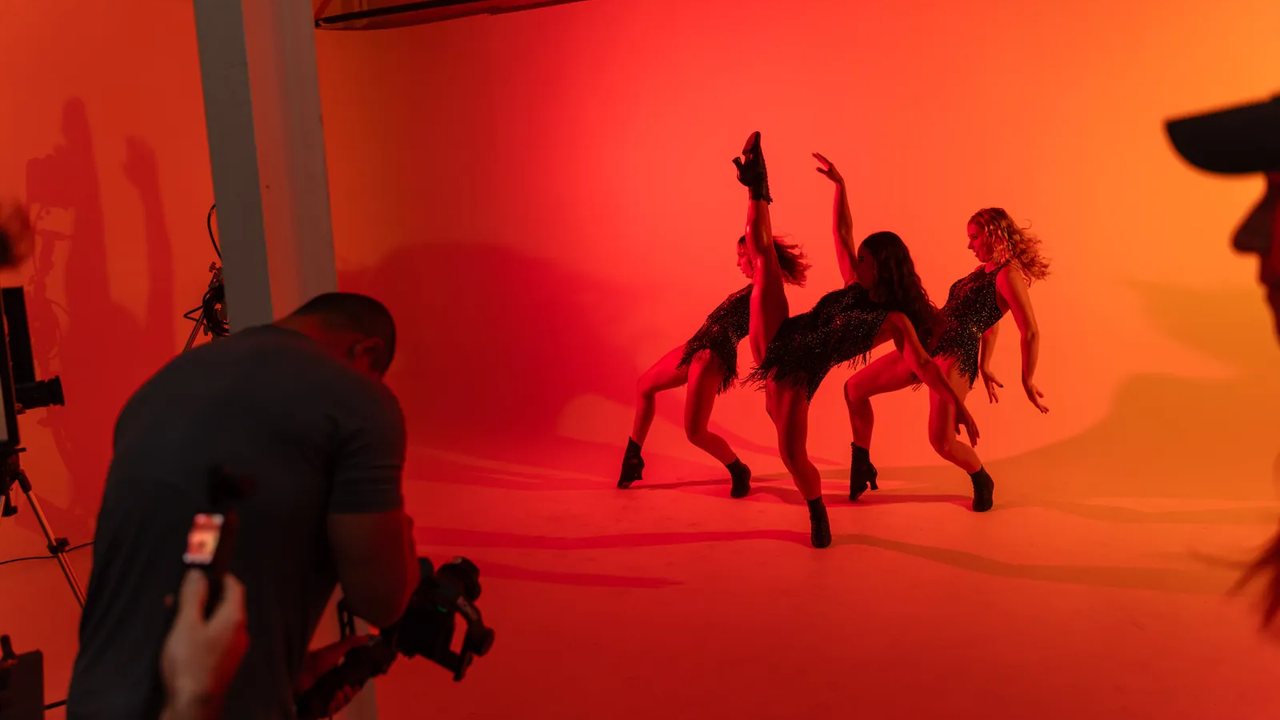 A camera operator filming three dancers against a red backdrop in the studio.